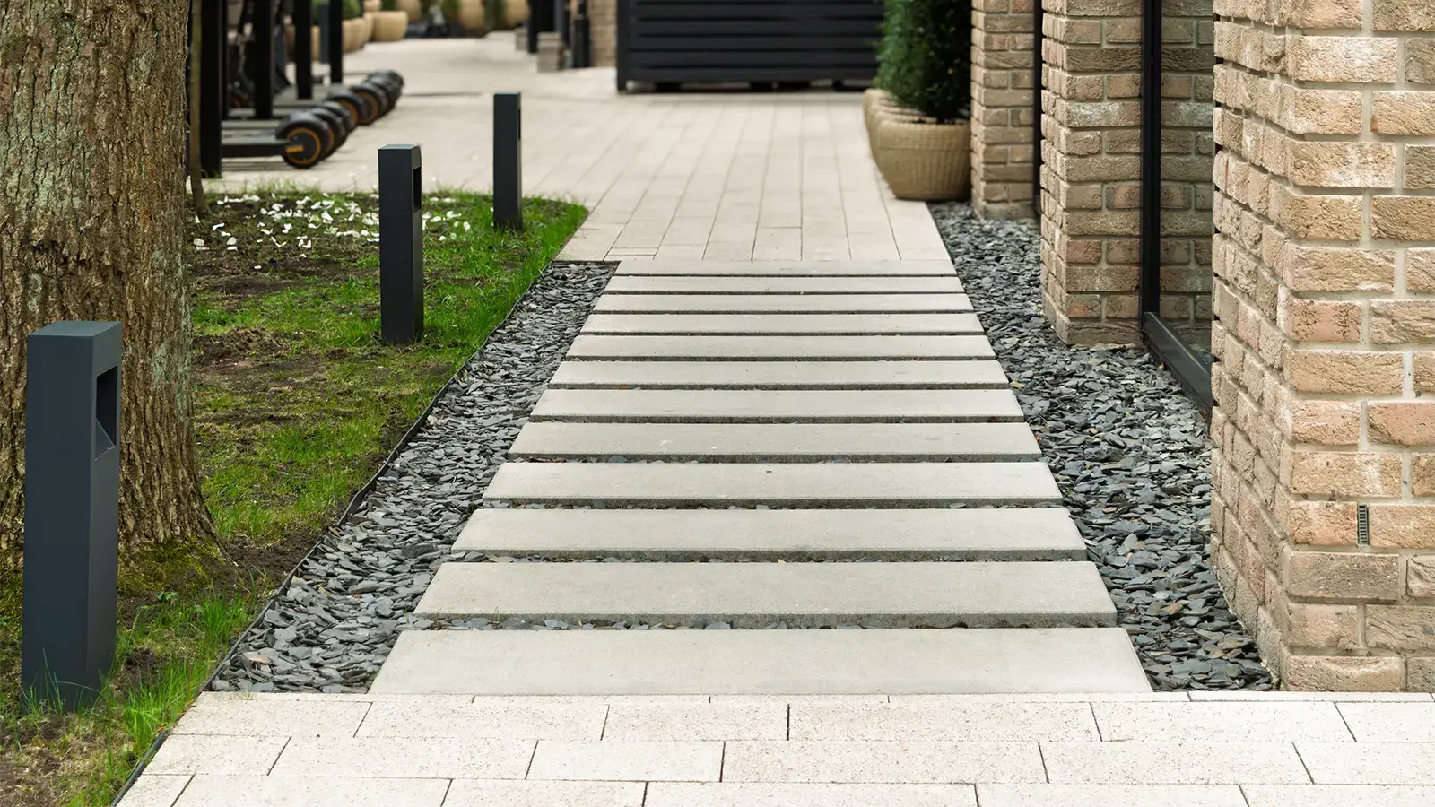Modern walkway made of concrete slabs leads along landscaped area with decorative chat rock and brick building, featuring outdoor lighting. 