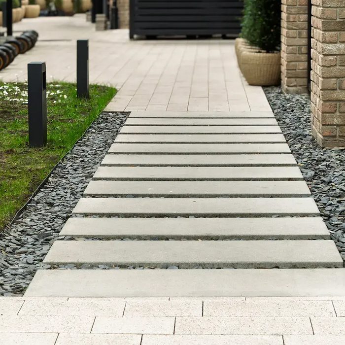 Modern walkway made of concrete slabs leads along landscaped area with decorative chat rock and brick building, featuring outdoor lighting. 