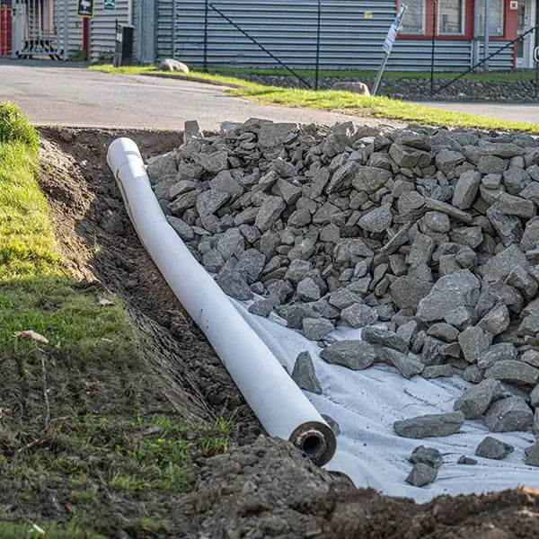 A white drainage pipe laid in a trench next to a large pile of gray rocks and a white landscape fabric, with grass and a building visible in the background.
