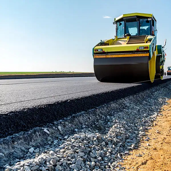 A yellow road roller compacts fresh asphalt on an outdoor highway under a clear blue sky.